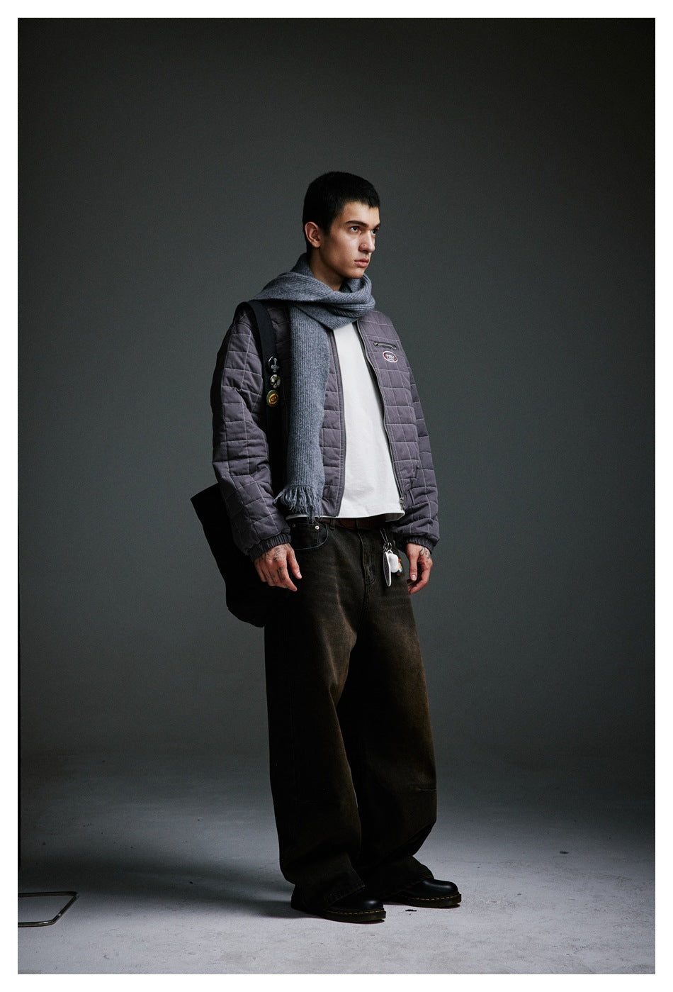 Model wearing a charcoal-gray quilted jacket with a gray scarf, white shirt, and dark wide-leg trousers.