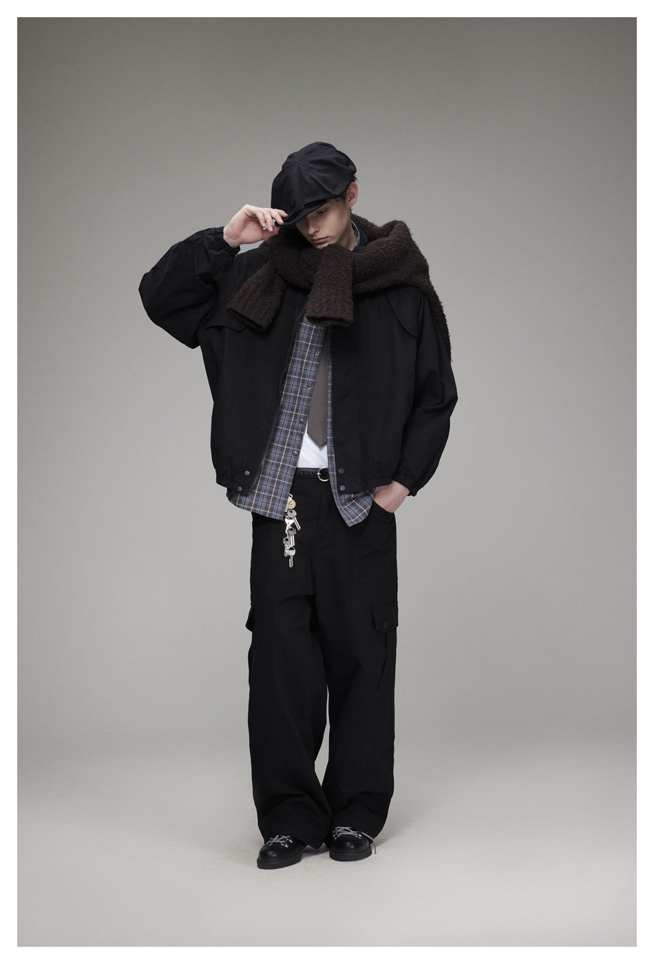 Model wears a black jacket, plaid shirt, brown scarf, black cargo trousers and cap, posing with one hand at the brim.