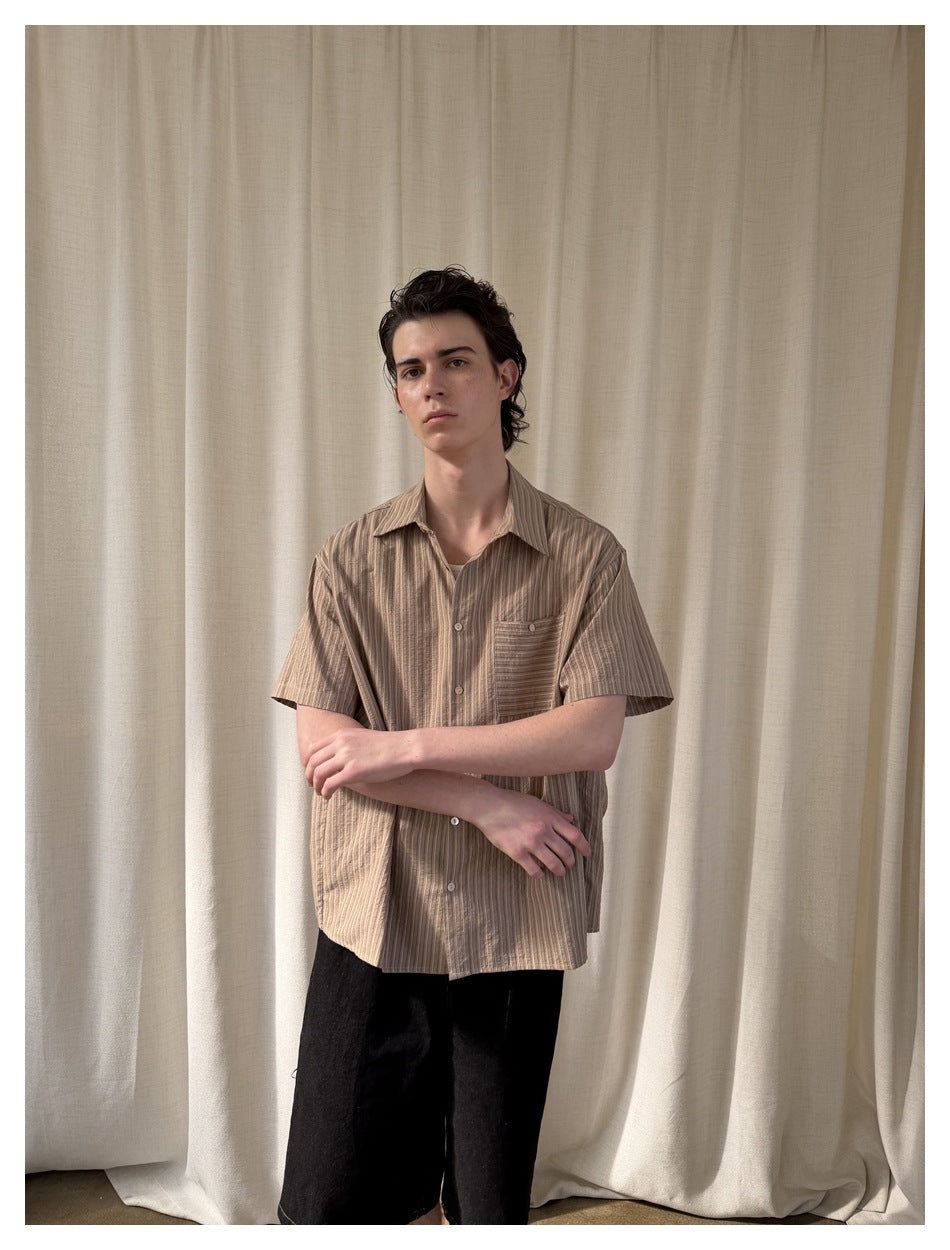 Model wearing a tan vertically striped short-sleeve button-up shirt with a chest pocket and black trousers.