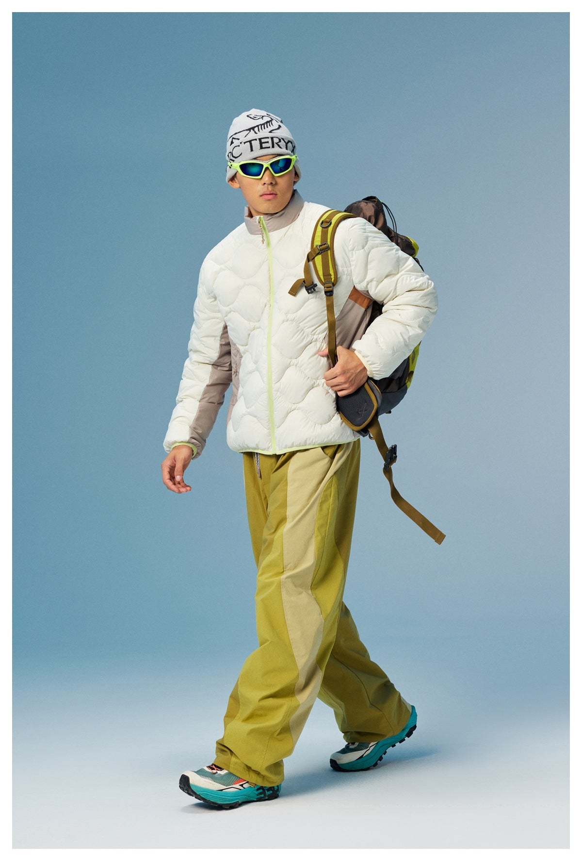 Model wearing a white wave-quilted jacket with taupe panels and lime zipper, olive trousers, and a backpack.