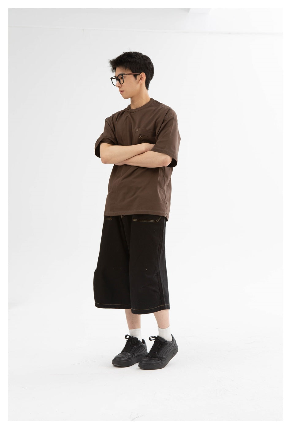 Model wearing a brown short-sleeve tee with a buttoned chest pocket and black wide-leg shorts, arms crossed.