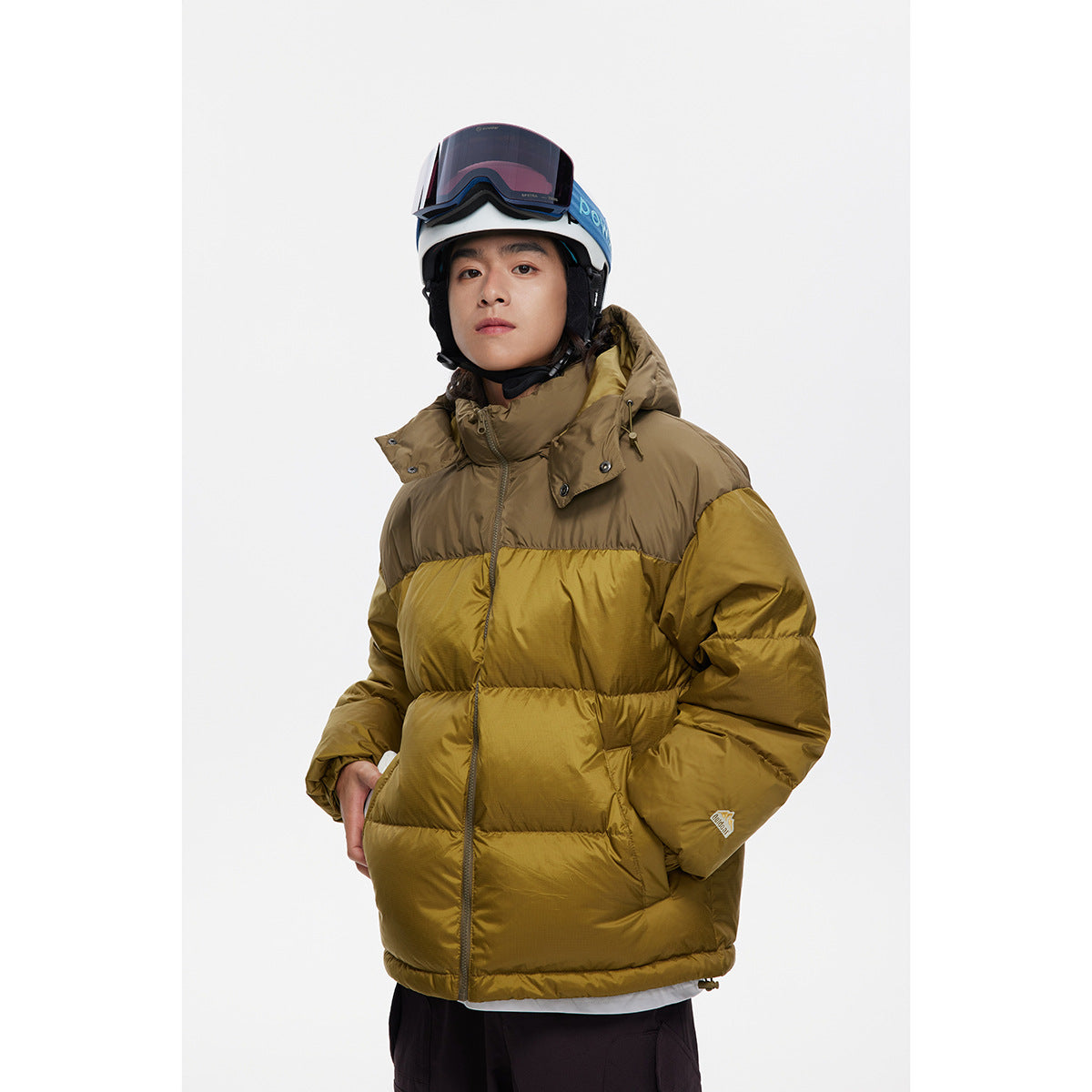 Front view of a model wearing an olive-and-mustard color-blocked hooded puffer jacket and ski helmet.