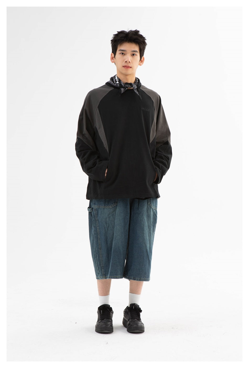 Full-length front view of a model wearing a black sweatshirt with charcoal panels and loose blue denim shorts.