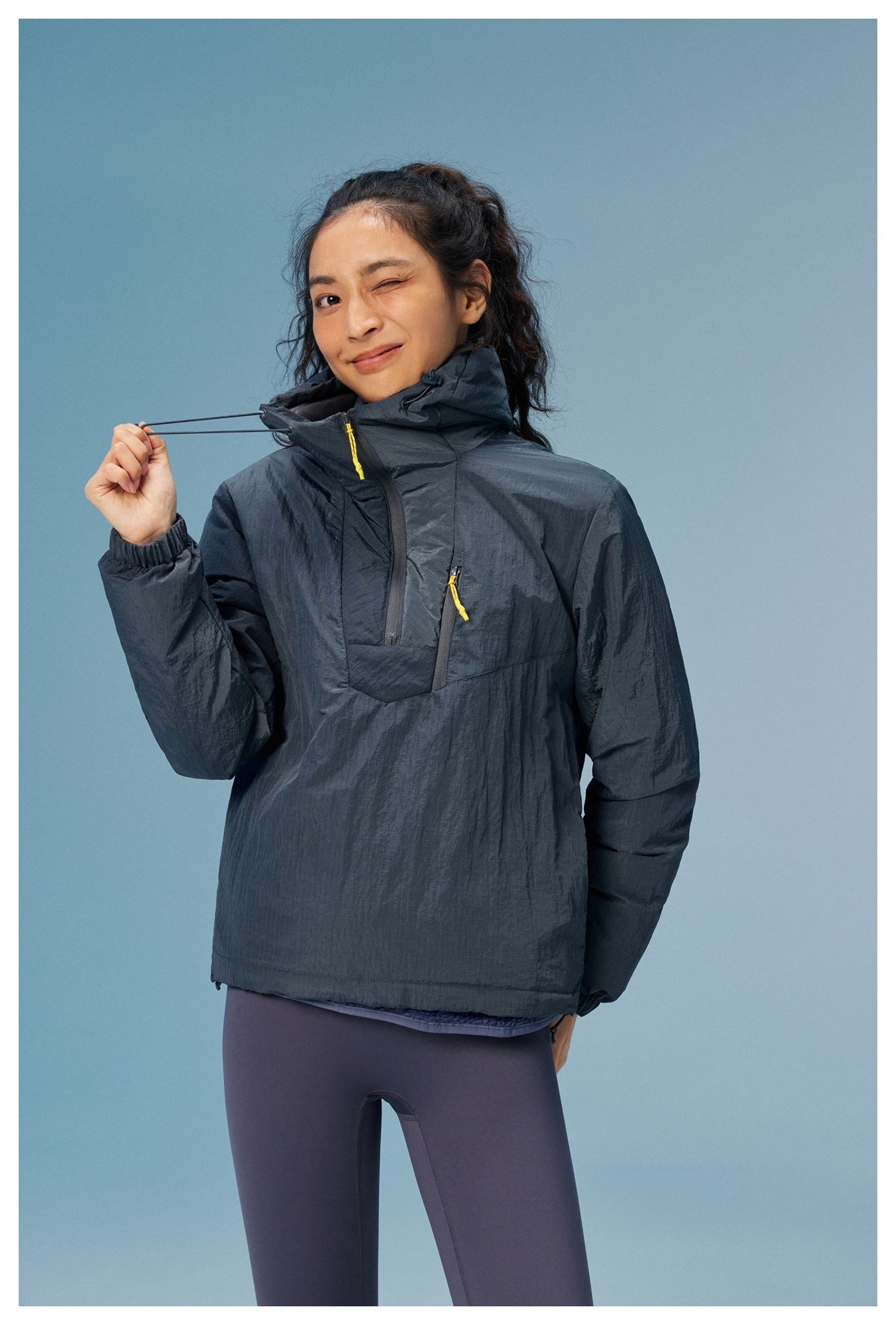 Woman wearing a dark gray hooded half-zip jacket with yellow zipper pulls, holding the hood drawcord