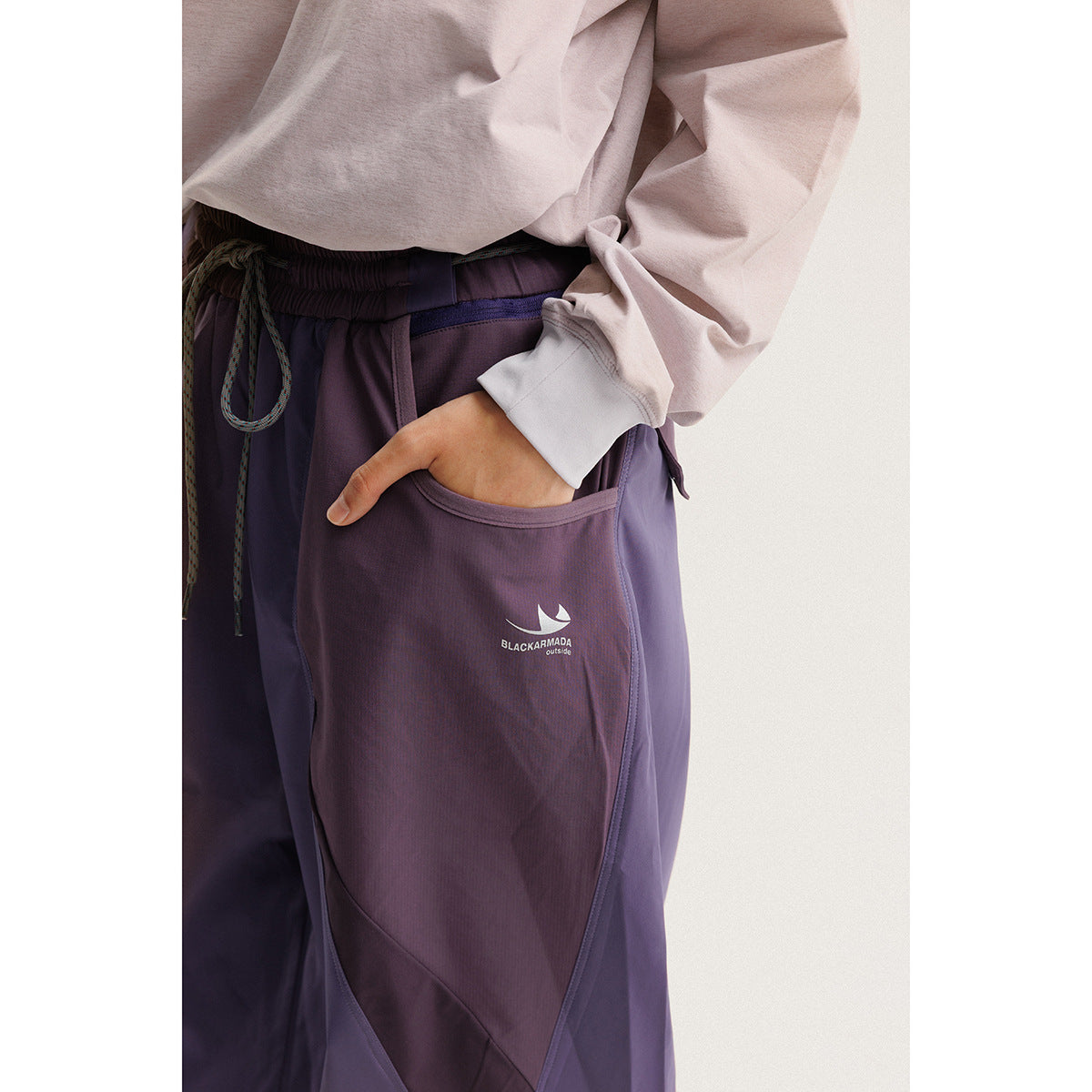 Close-up of purple outdoor pants showing a curved pocket, drawstring waist, panel seams and a small white logo.