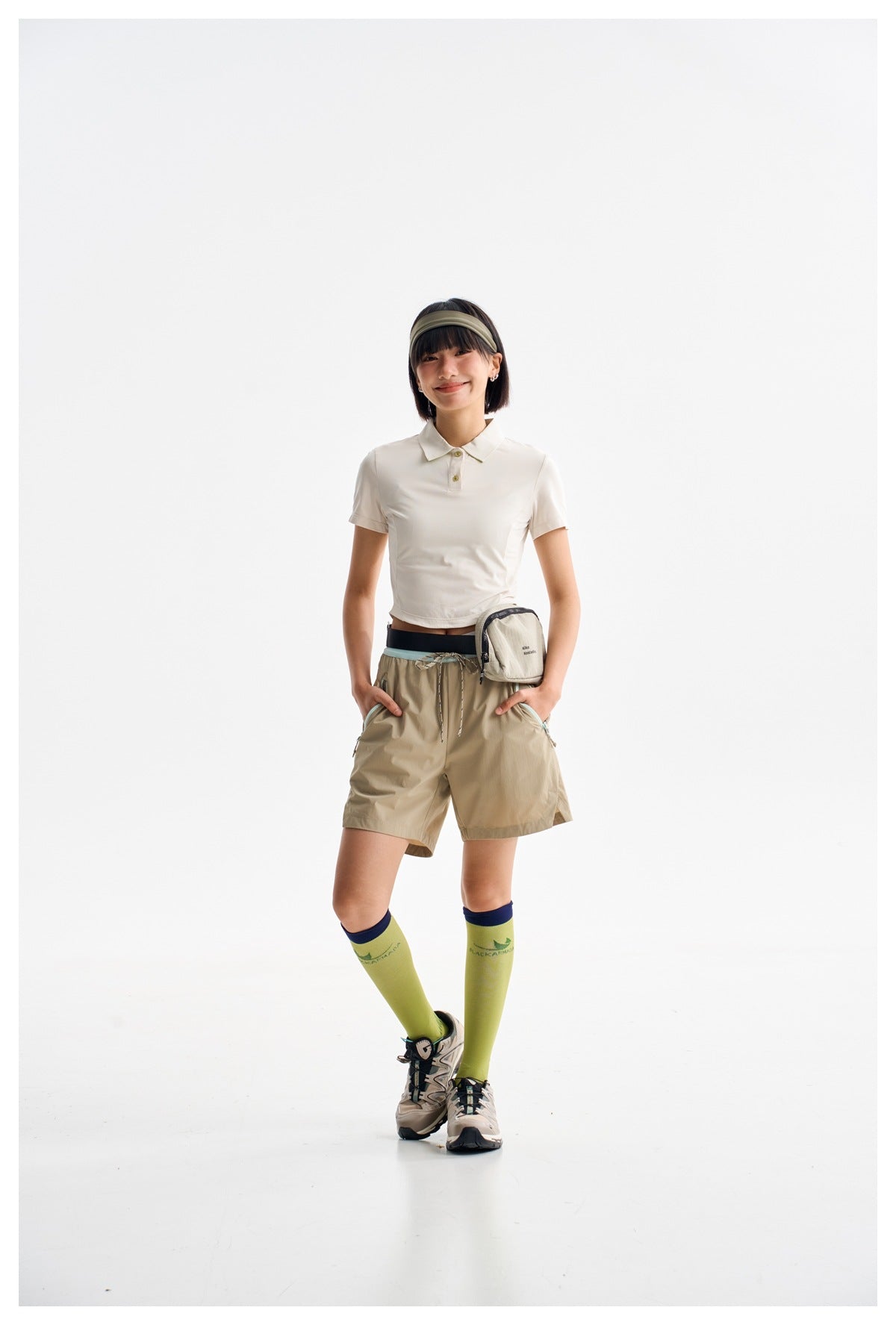 Model wearing a white cropped polo, khaki shorts, lime knee-high socks and hiking shoes against a white studio background.