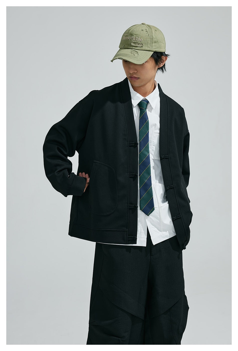 Model in black V-neck frog-button jacket with large round pocket over white shirt and striped tie, green cap
