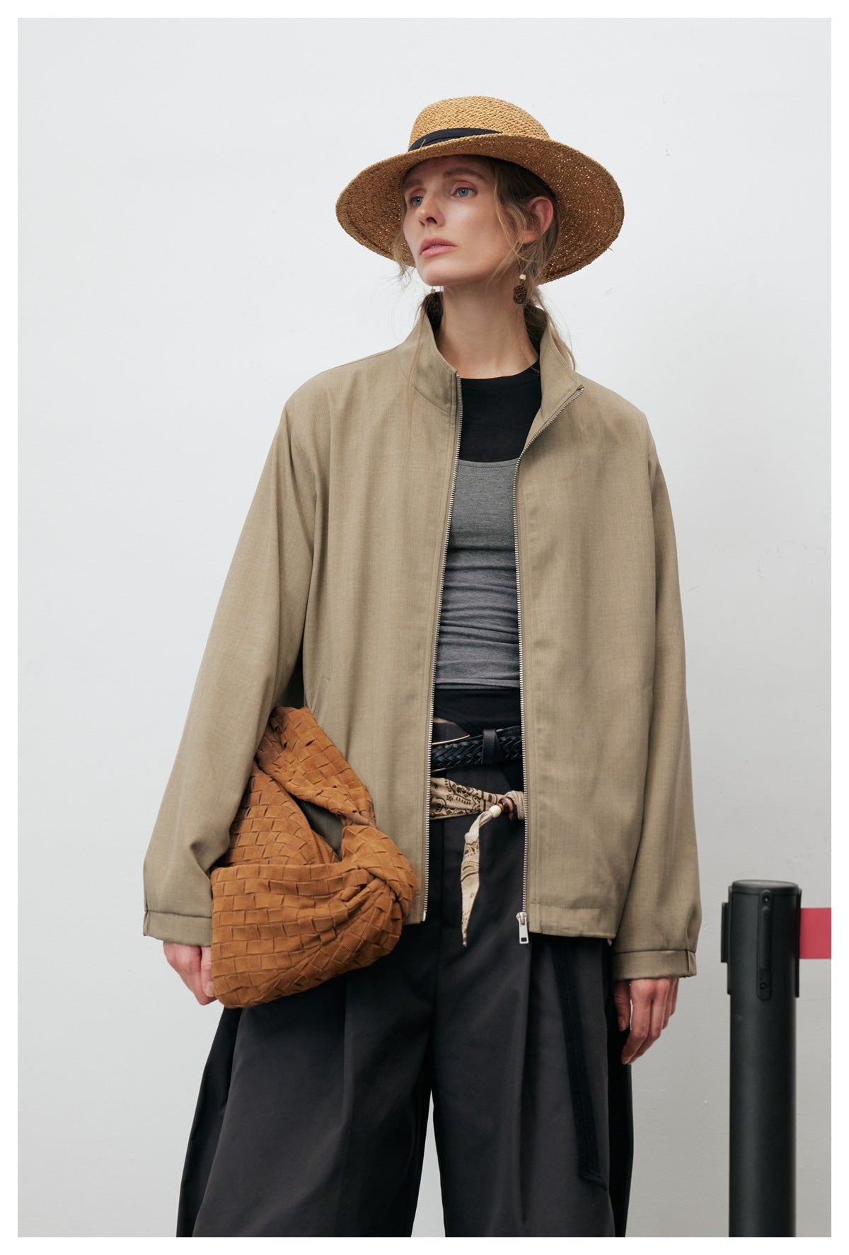 Close view of a model wearing a khaki stand-collar jacket open over a gray top with dark pants and a woven bag