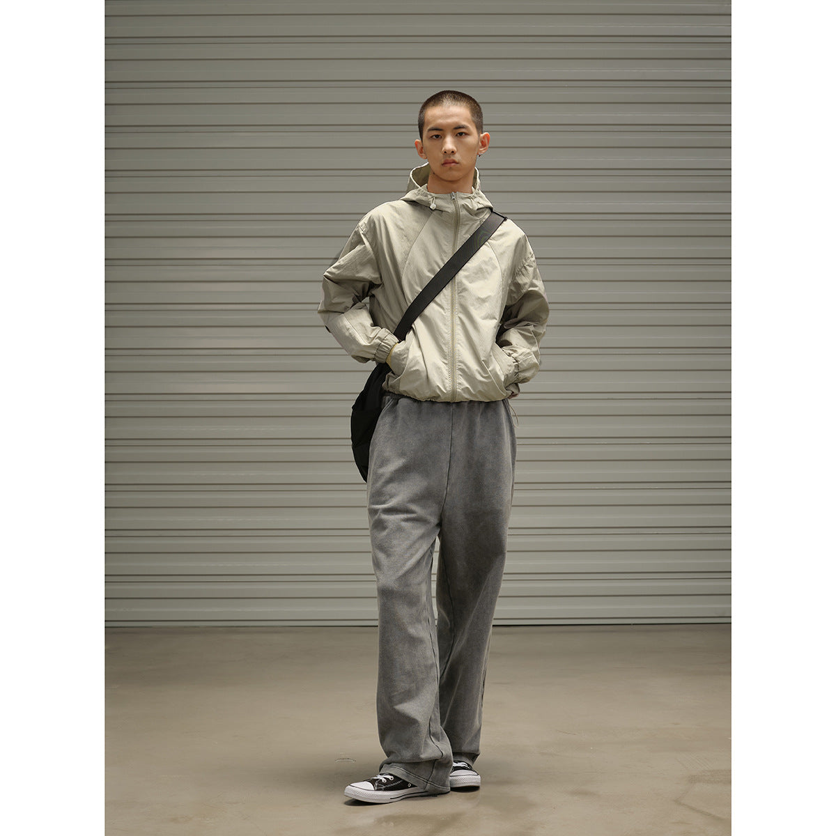 Model wearing a light beige hooded jacket and loose charcoal gray pants with a black crossbody bag.