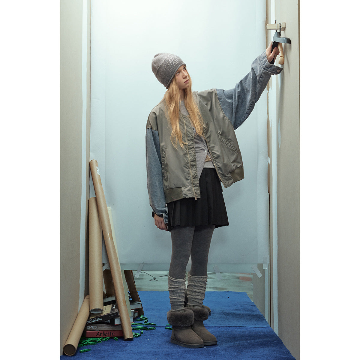 Model in a gray-green bomber jacket with blue denim sleeves, shown standing with one arm raised.