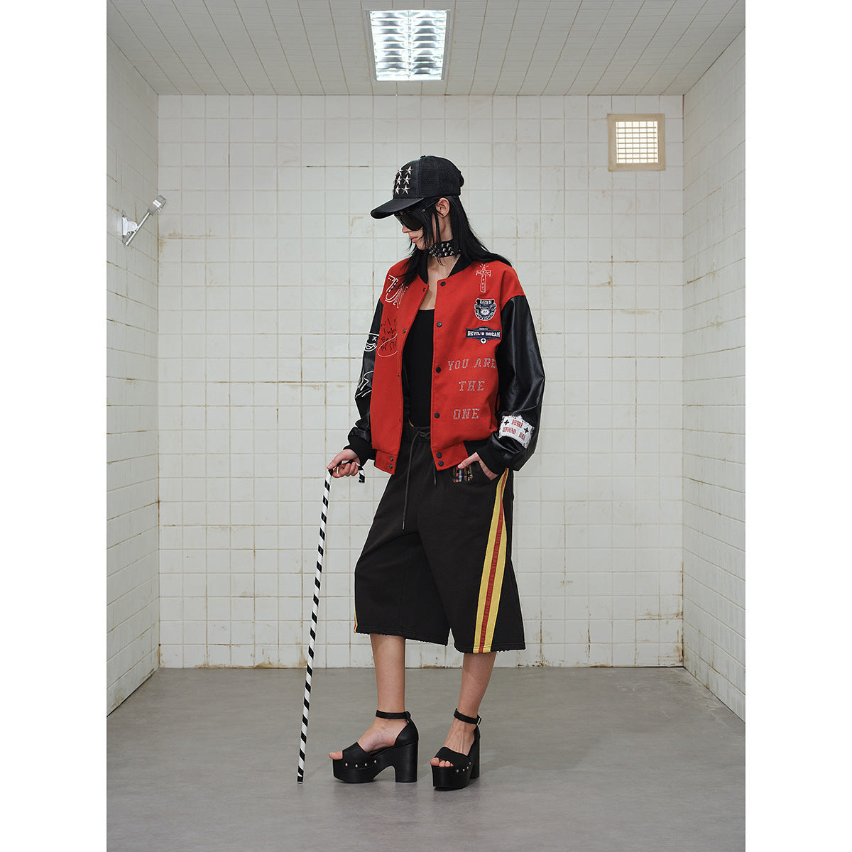 Model in a red varsity jacket with black sleeves and a black cap, paired with black shorts with side stripes.