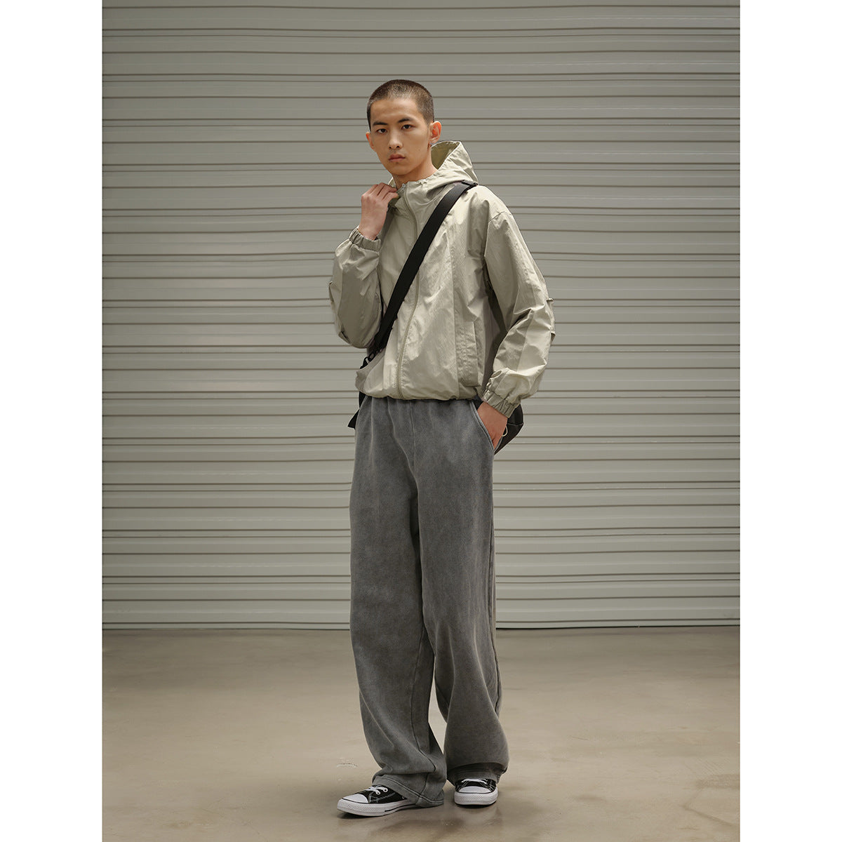 Model in a light beige hooded jacket and loose charcoal gray pants, posing at an angle with a black crossbody bag.