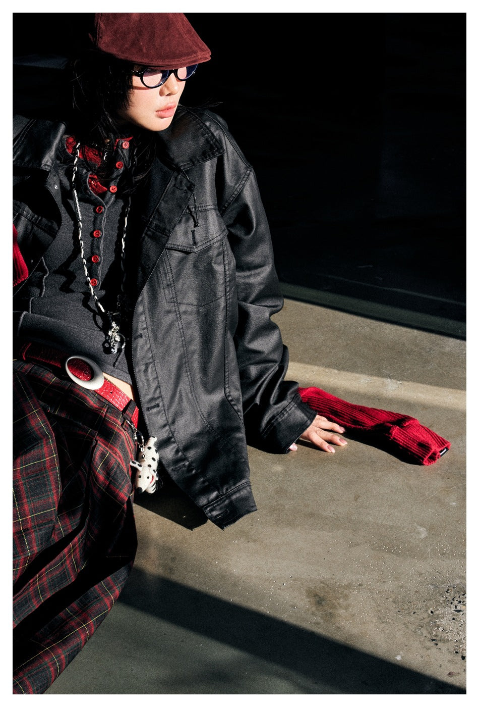 Seated model in an oversized black jacket with flap pockets, a red scarf, and a red-and-black plaid skirt.