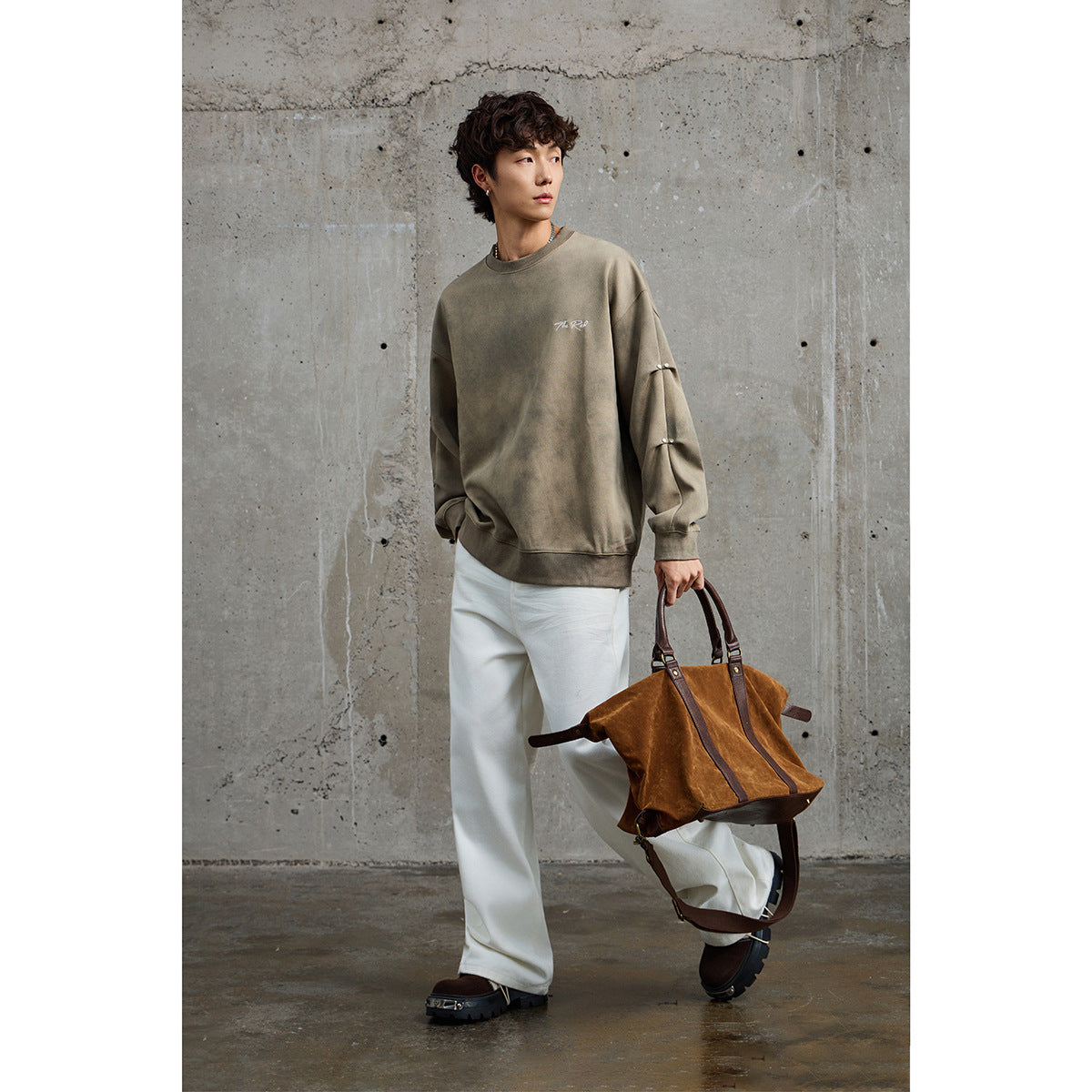 Model wearing a taupe oversized sweatshirt with white chest script and sleeve studs, paired with white trousers.