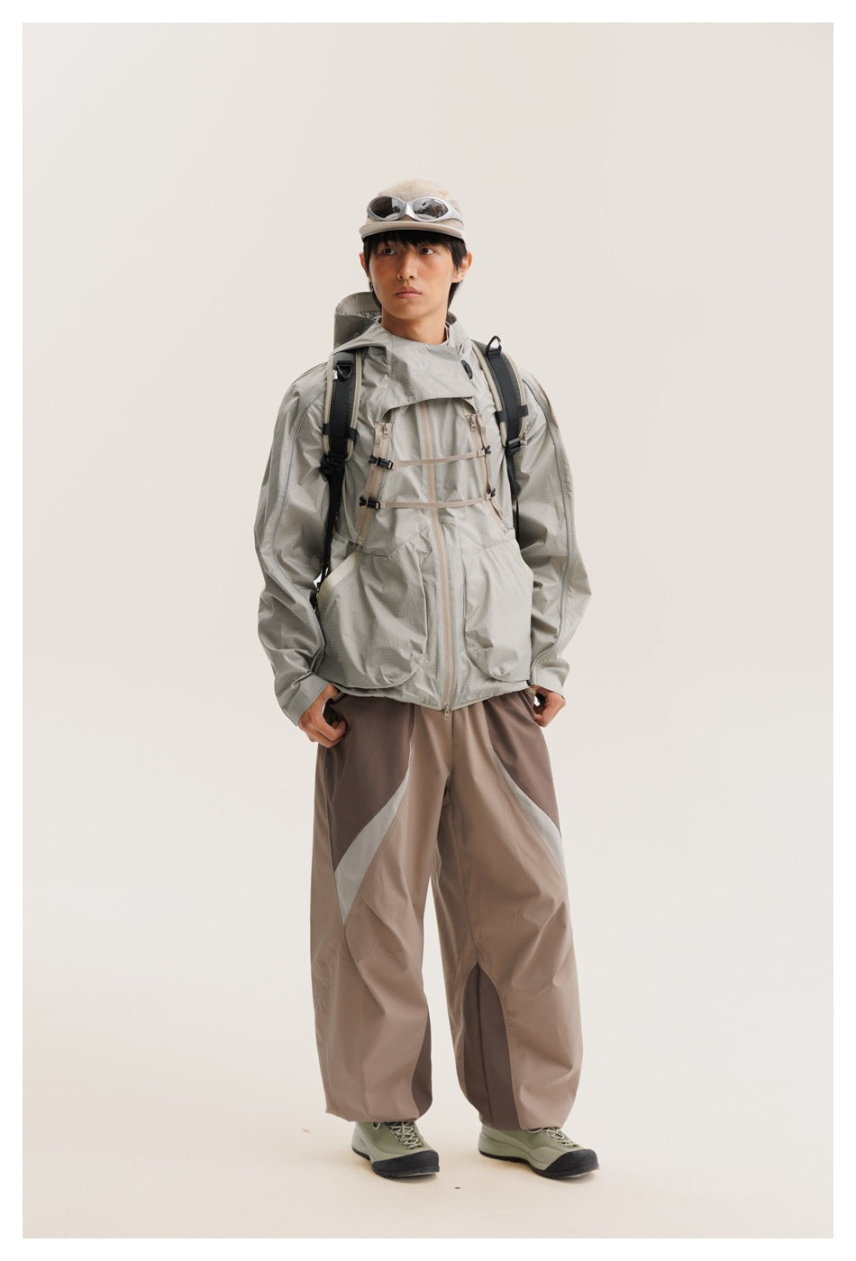 Model wearing a light gray hooded jacket with chest straps and large pockets, two-tone taupe pants and a backpack.