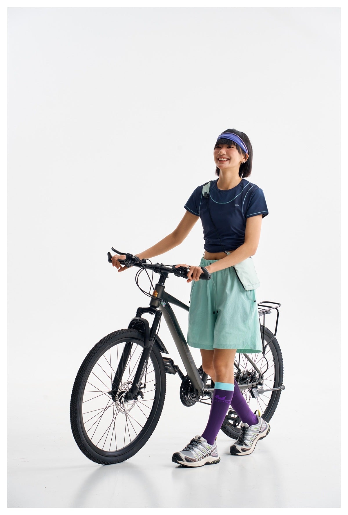 Model in a navy piped crop top and mint shorts stands with a bicycle, wearing purple socks and a blue headband.