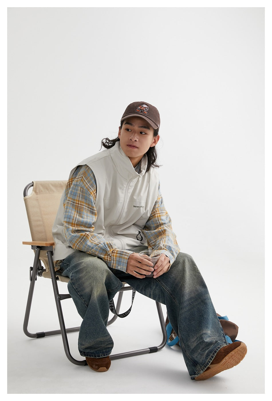 Seated model wearing an off-white sleeveless zip vest over a blue and ochre plaid shirt.