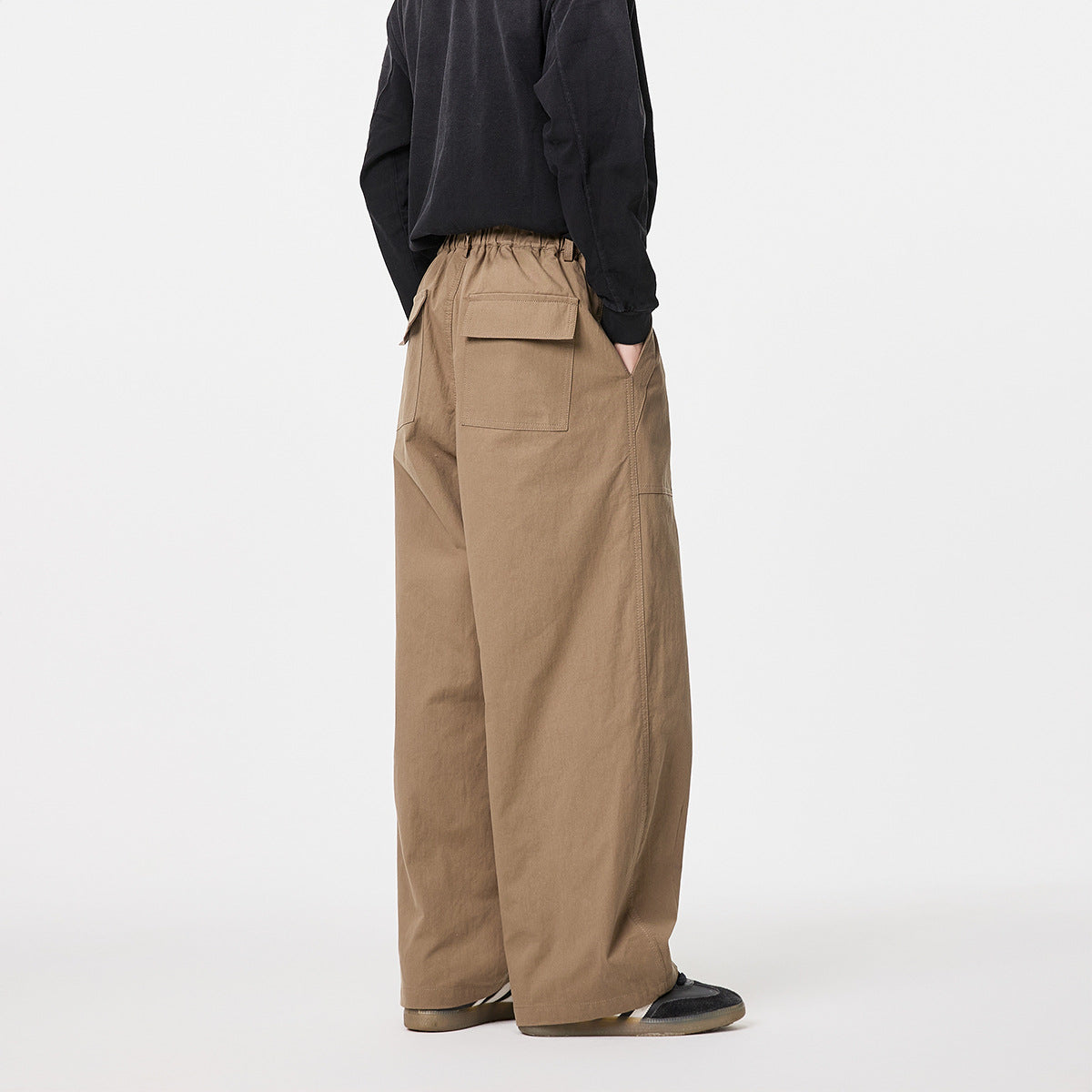 Rear three-quarter view of a model wearing tan wide-leg trousers with an elastic waistband and flap back pockets.