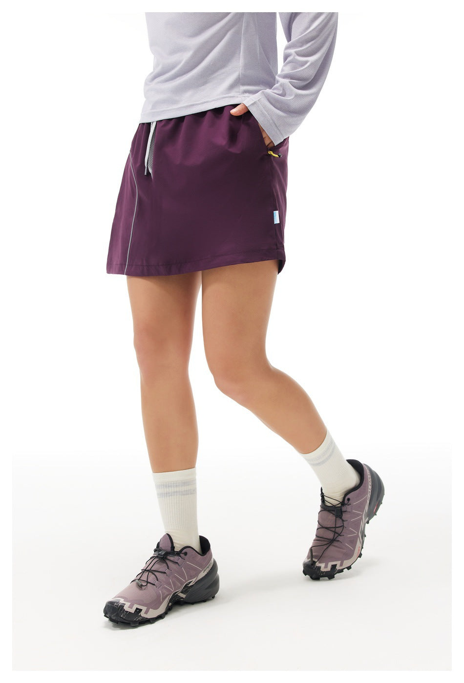 Side view of a woman wearing a purple skort with hand in the side pocket, lavender top, white socks and trail shoes