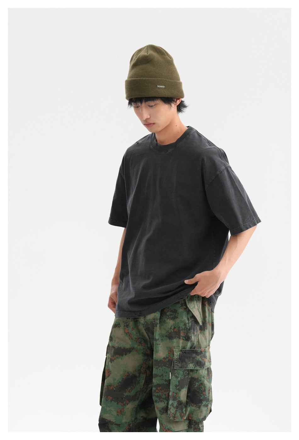 Closer view of a model in an oversized charcoal T-shirt and green camouflage cargo pants with an olive beanie.