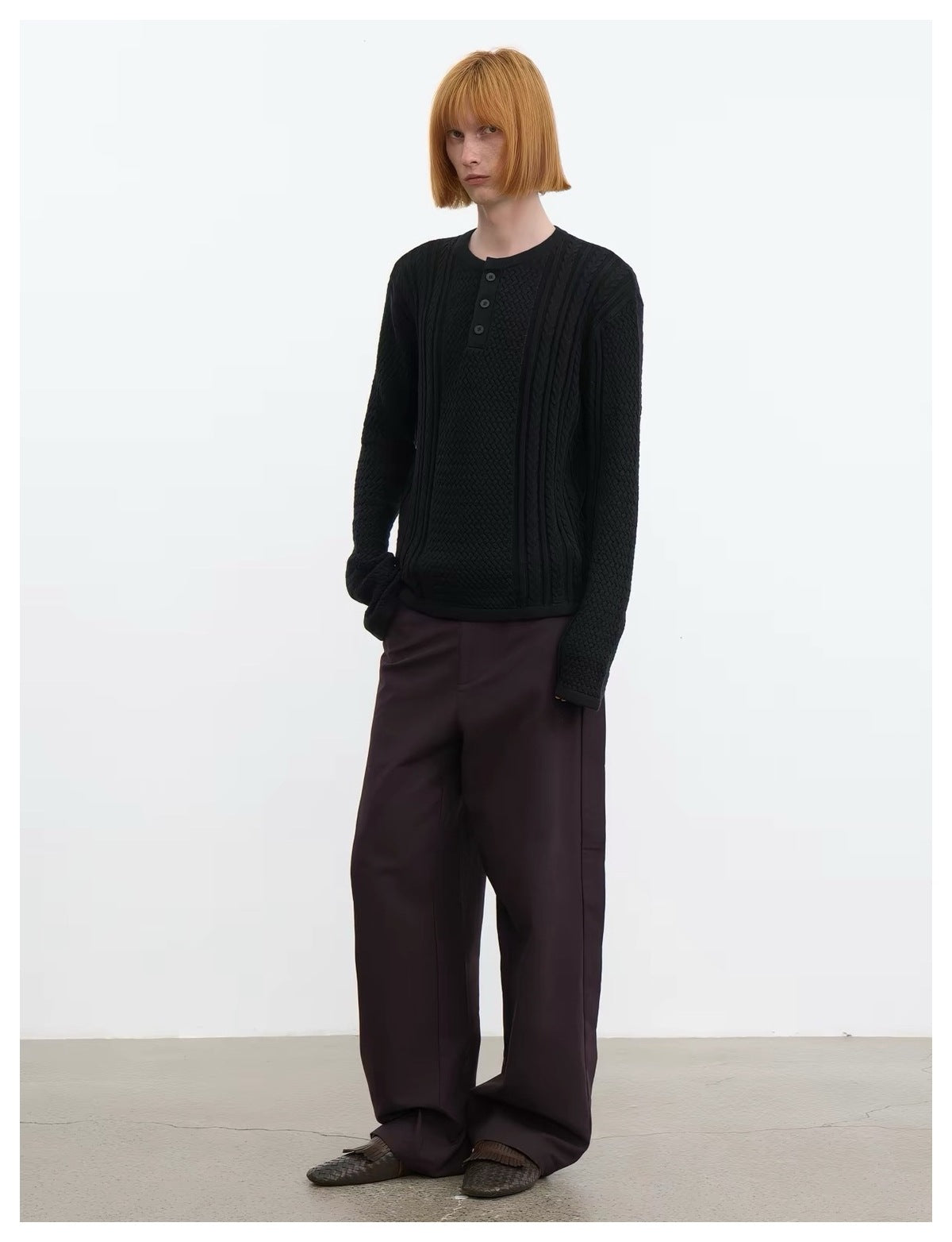 Model in black cable-knit Henley sweater and brown wide-leg trousers, angled stance with hand in pocket