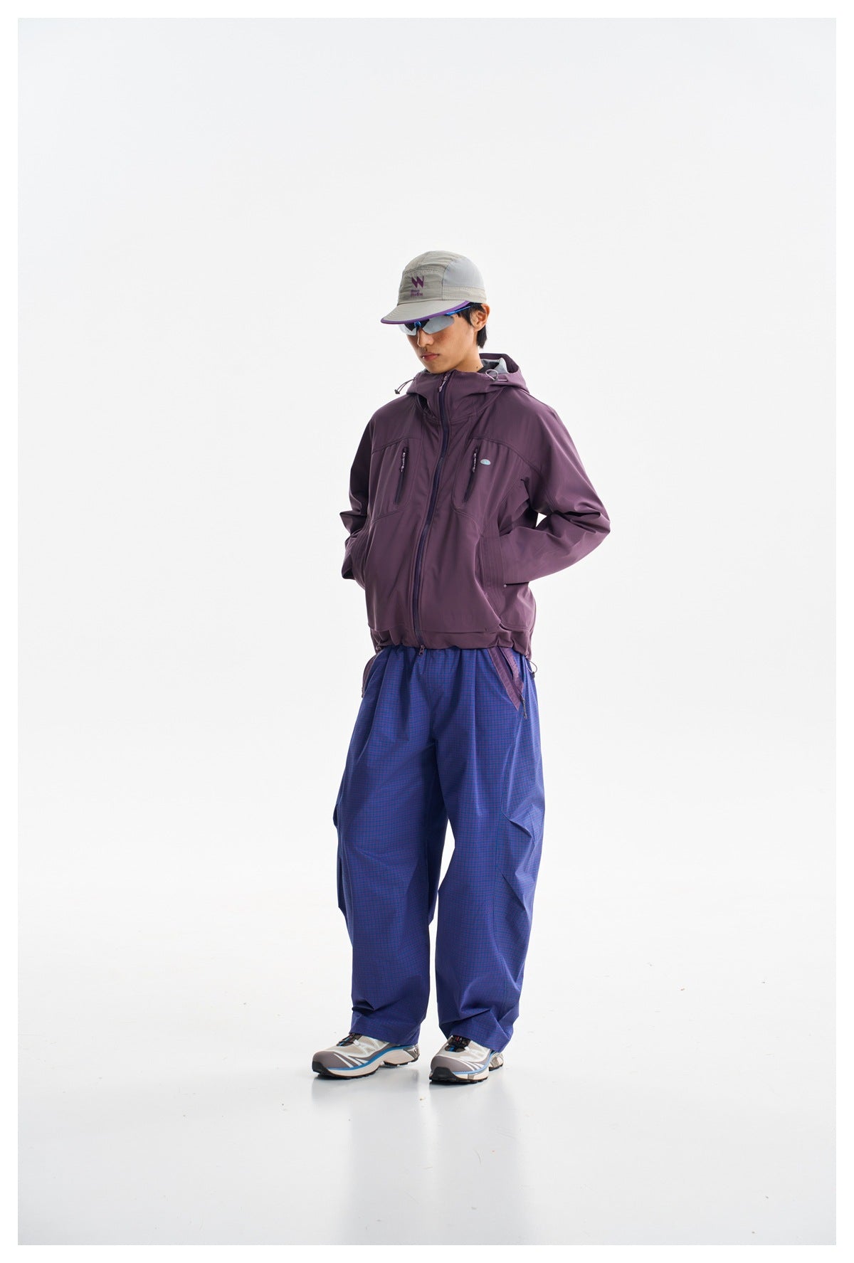 Model wearing a plum hooded jacket with zip pockets and loose blue checked trousers, shown full length.
