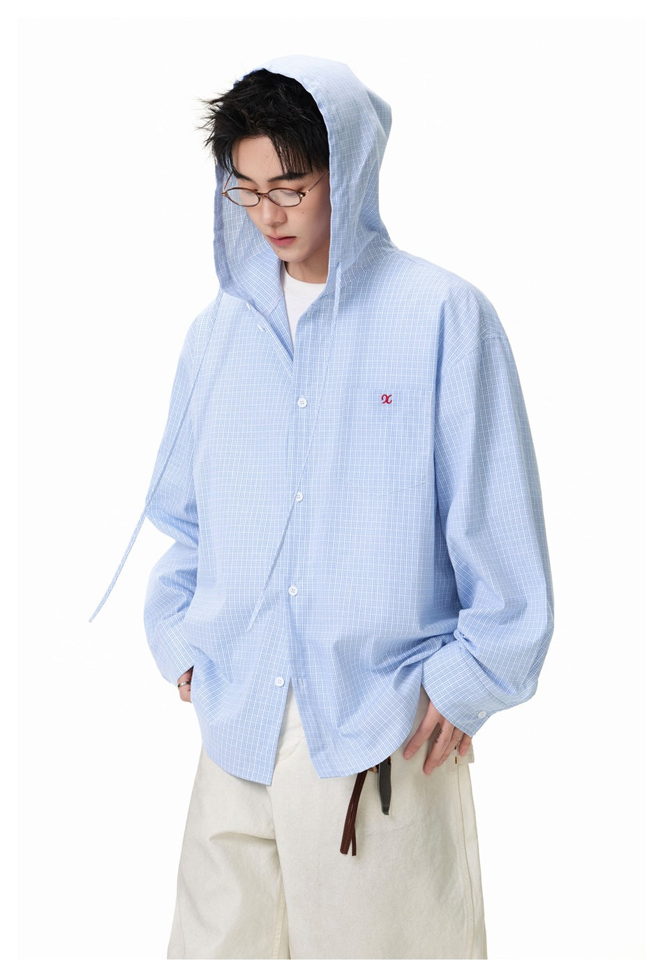 Model wears the hood up on an oversized light blue checked button-up shirt with a chest pocket and cream trousers.