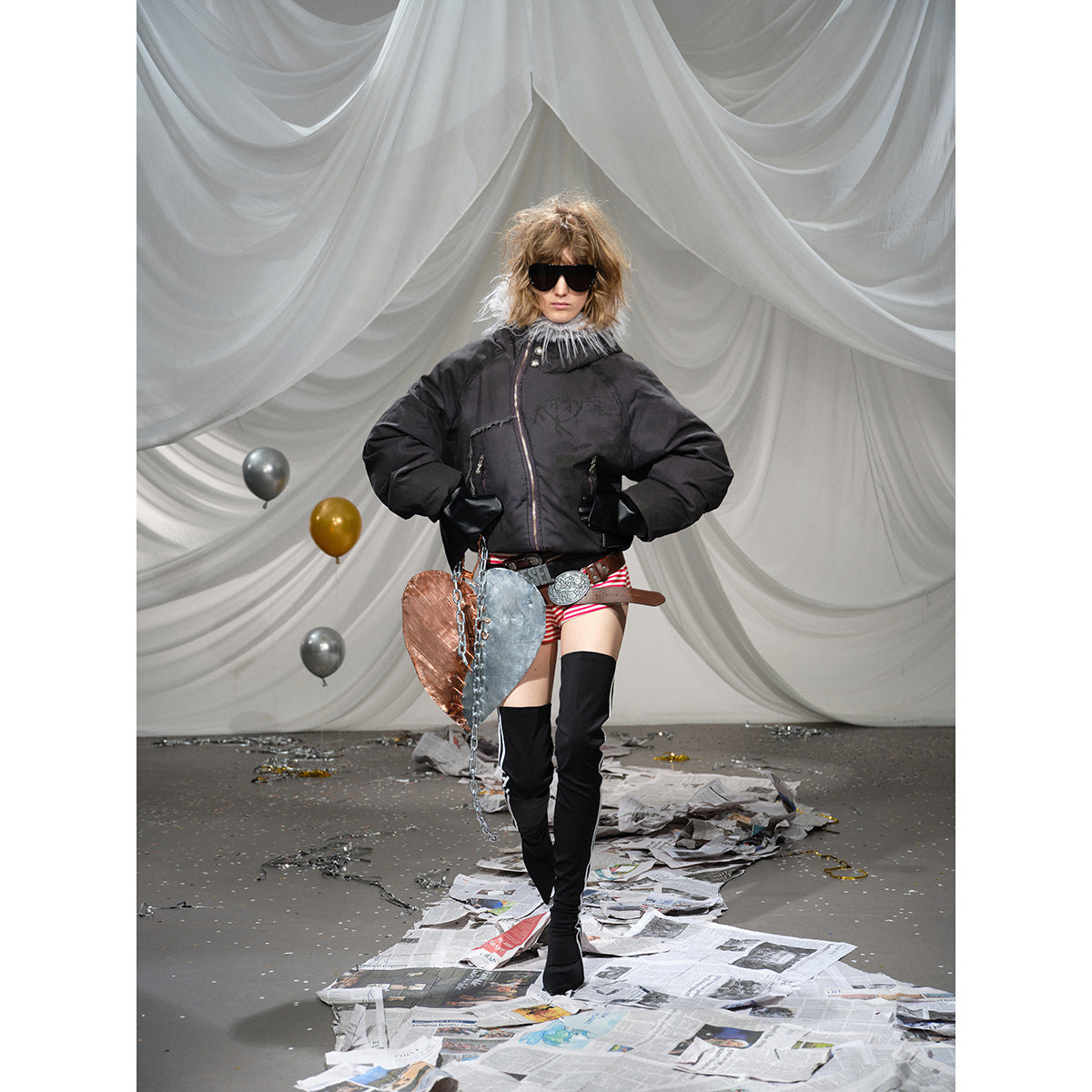 Model wearing a black padded jacket with a gray fur-trimmed hood, red-and-white striped shorts and black thigh-high boots.