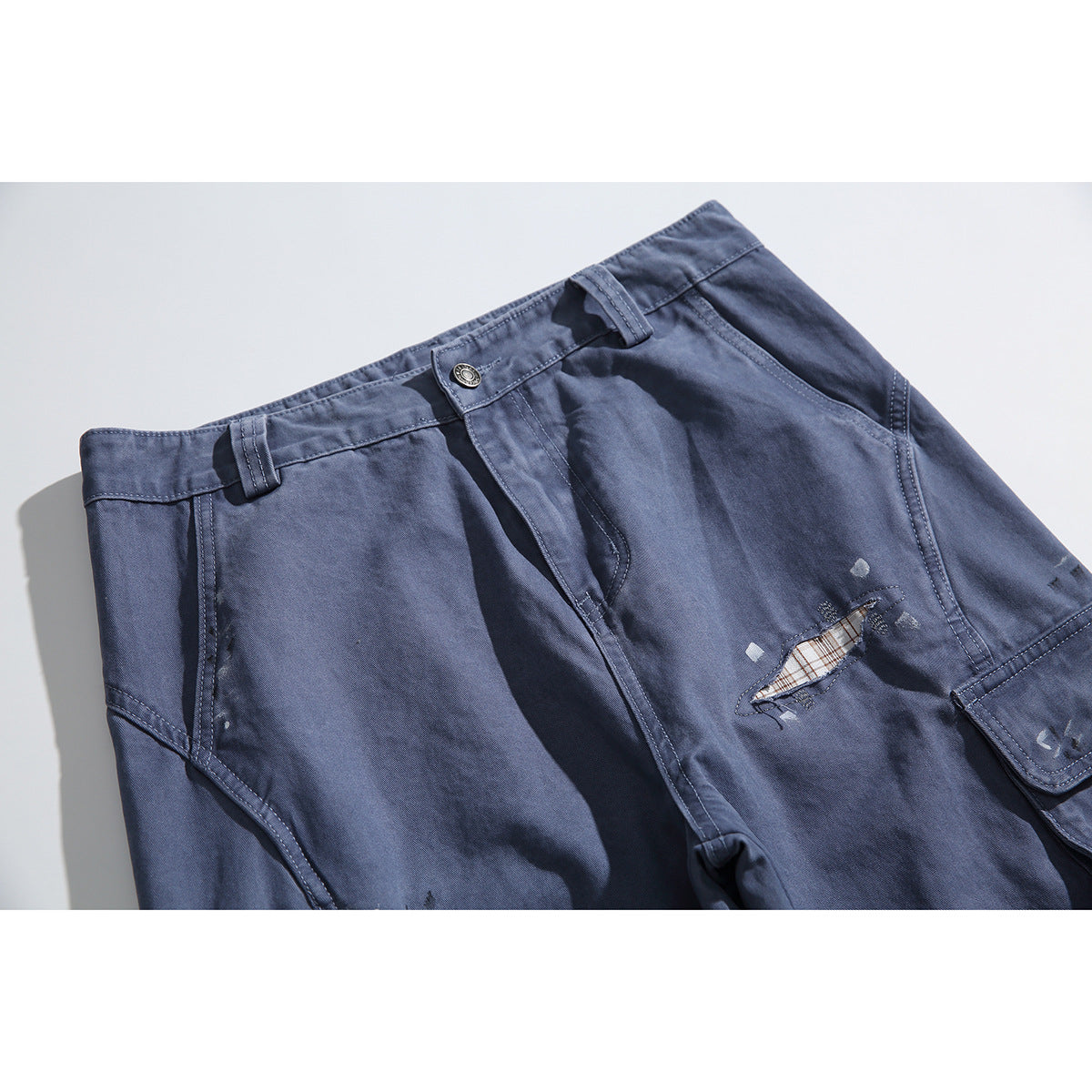 Close-up of blue-gray cargo pants showing the waistband, fly, pocket seams, and a distressed opening with plaid fabric
