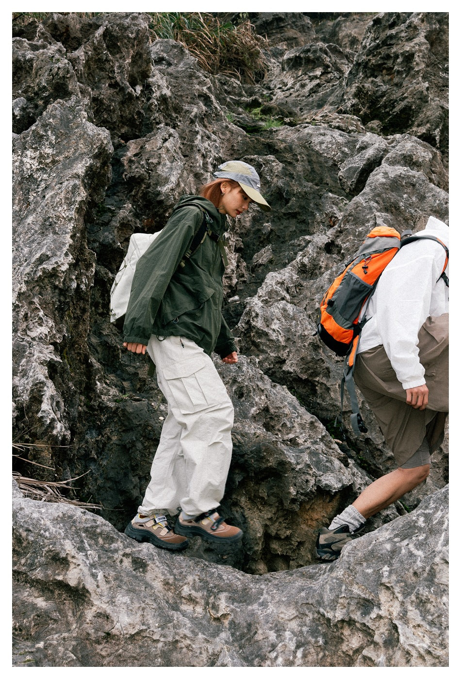 Person in a green hooded jacket and light cargo pants climbing rocks with a second person in white