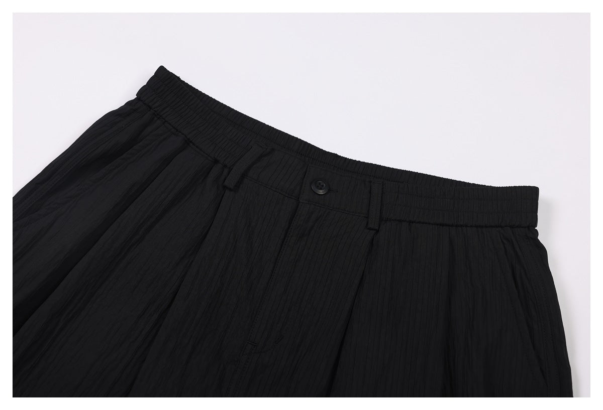 Close-up of black trousers showing the elasticated waistband, button, belt loop, and front pleats.