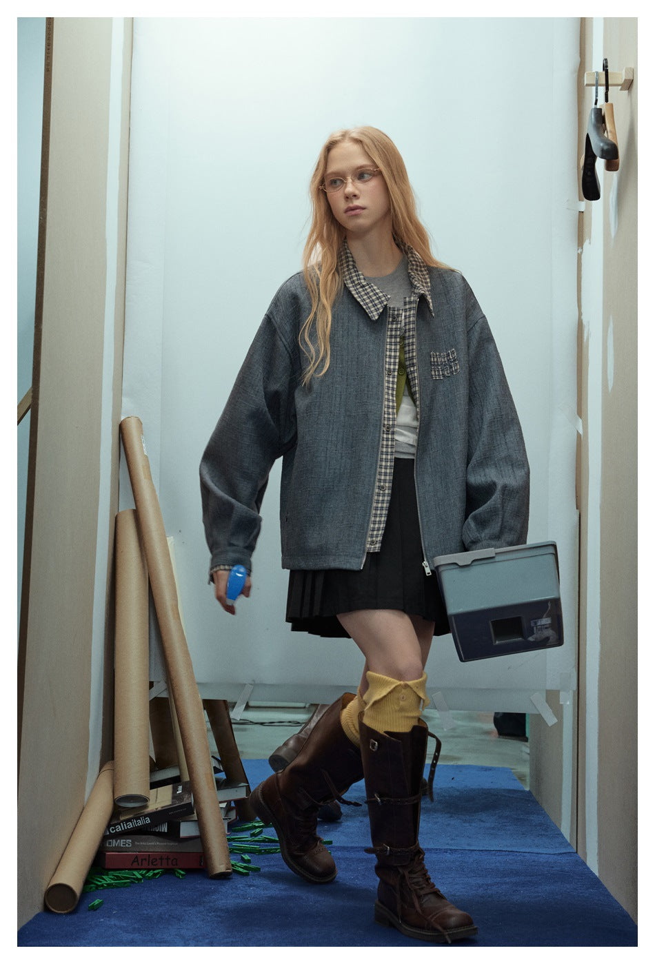 Model walking in an oversized blue-gray jacket with plaid collar and placket, a black skirt, and tall brown boots.