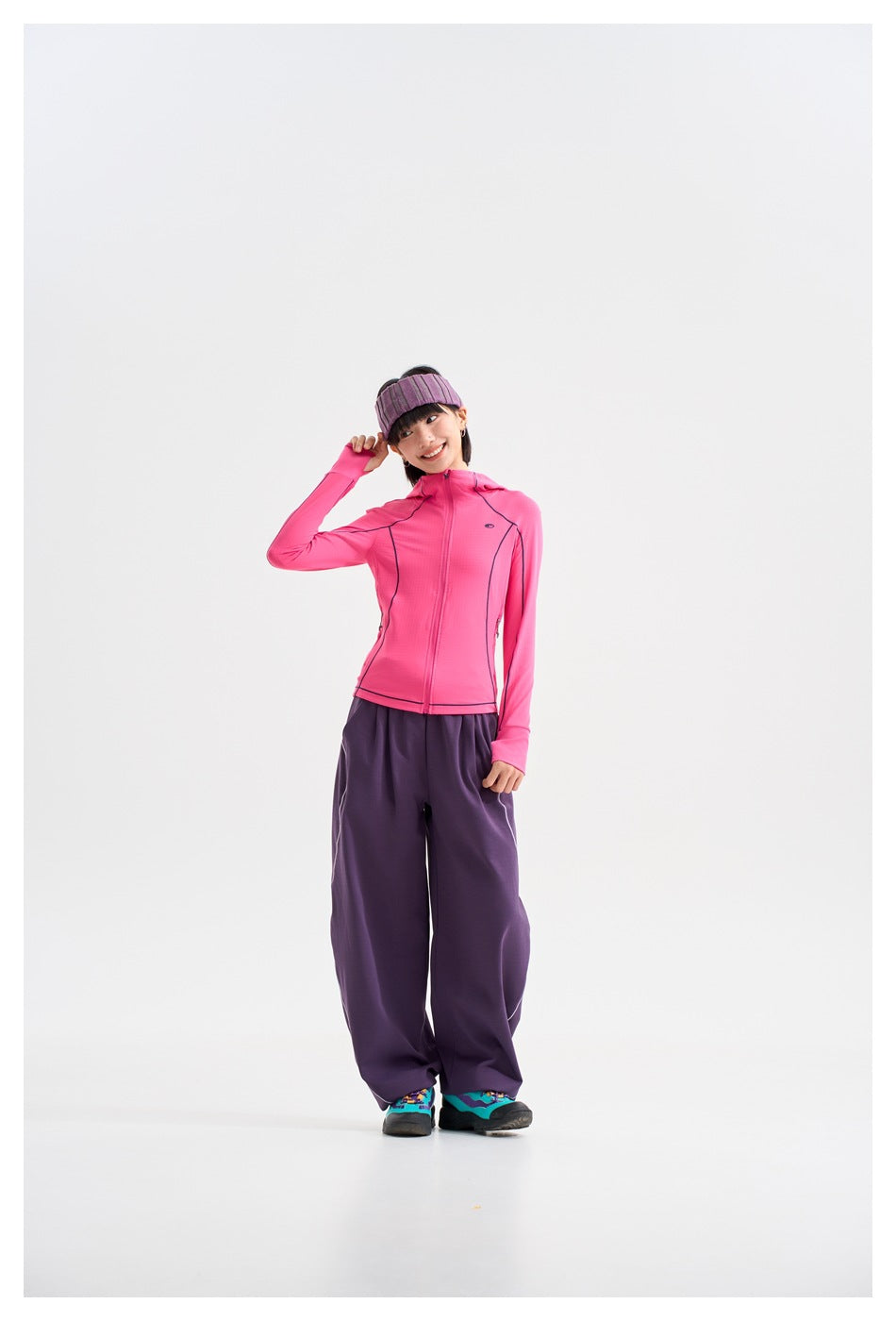 Model wearing a bright pink zip-front hooded jacket with dark piping and loose purple pants, posing with one hand at her