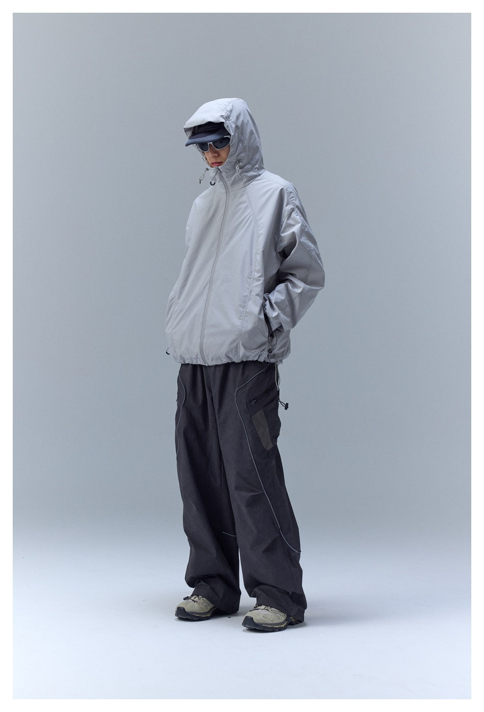 Model wearing a light gray hooded zip jacket and loose black cargo pants with curved piping.