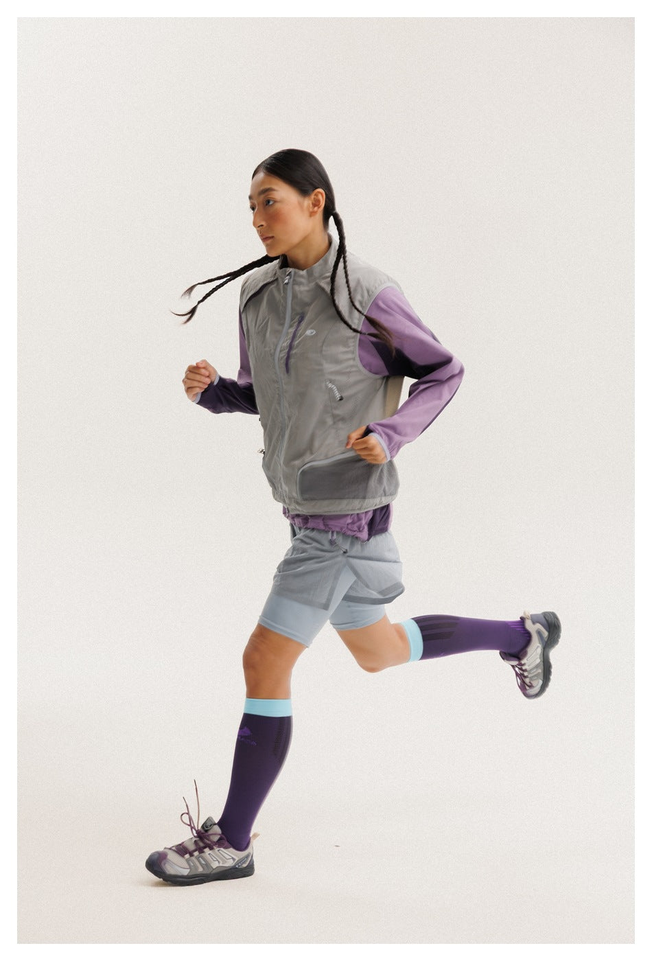 Model running in profile, wearing a gray zip vest, purple sleeves, gray shorts, and purple knee-high socks.