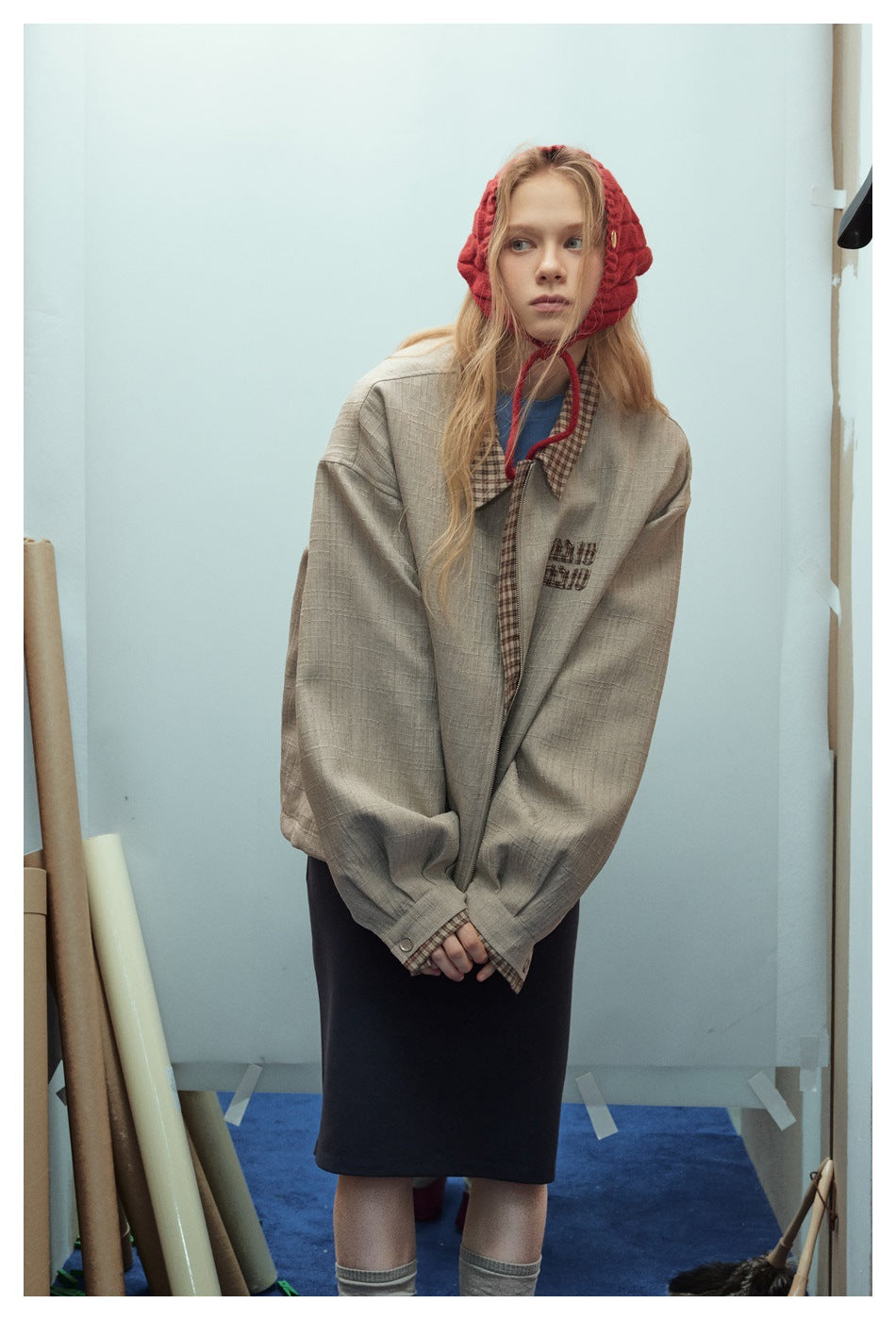 Model wearing an oversized taupe jacket with a checked collar and chest embroidery, a red knit bonnet and dark skirt.