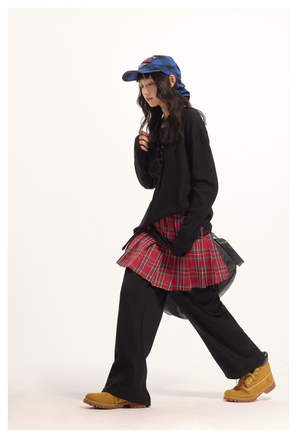 Model wearing a black lace-up top, red plaid pleated skirt over black trousers, and a blue patterned cap.