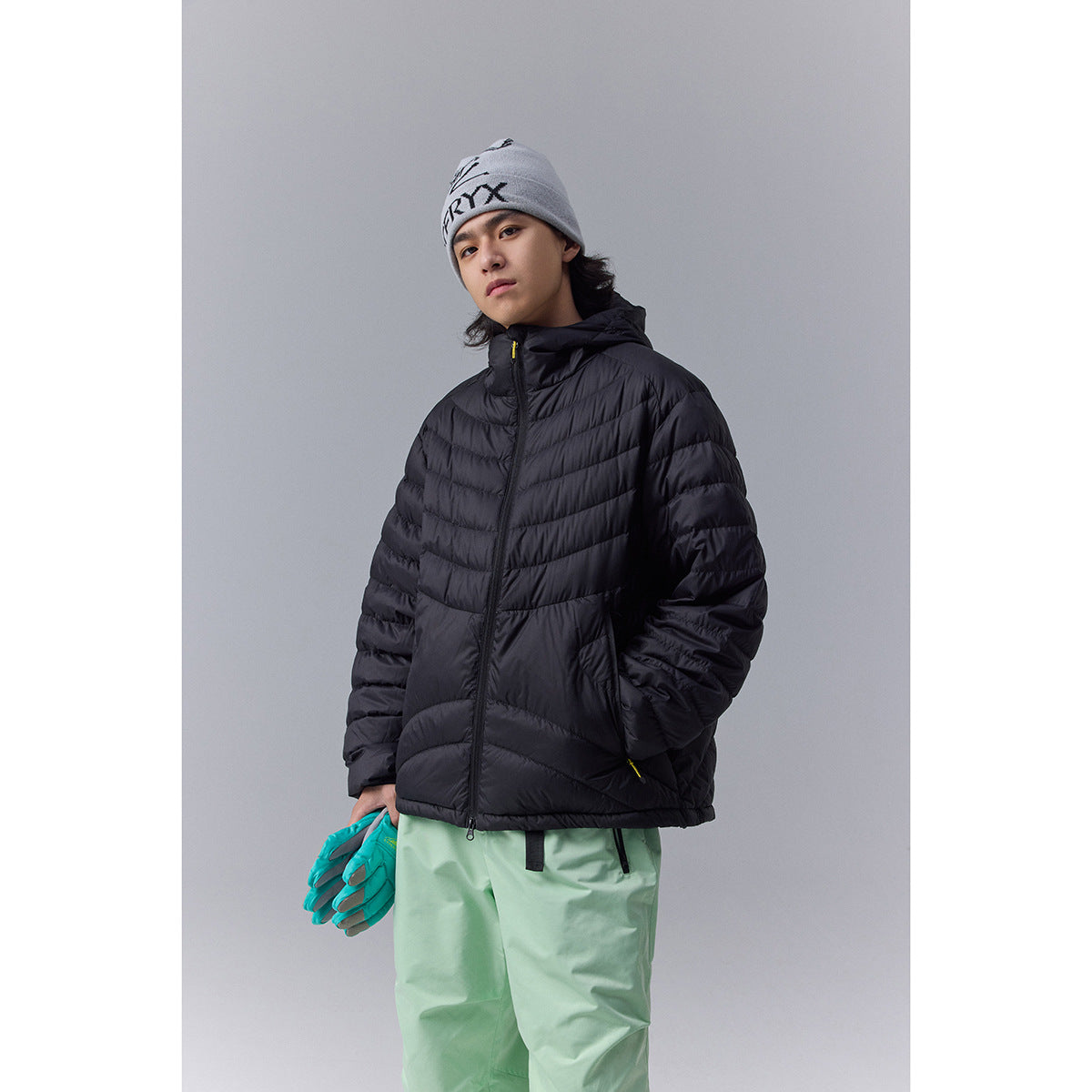 Model wearing a black hooded quilted jacket, gray beanie, and mint-green ski pants, holding teal gloves.
