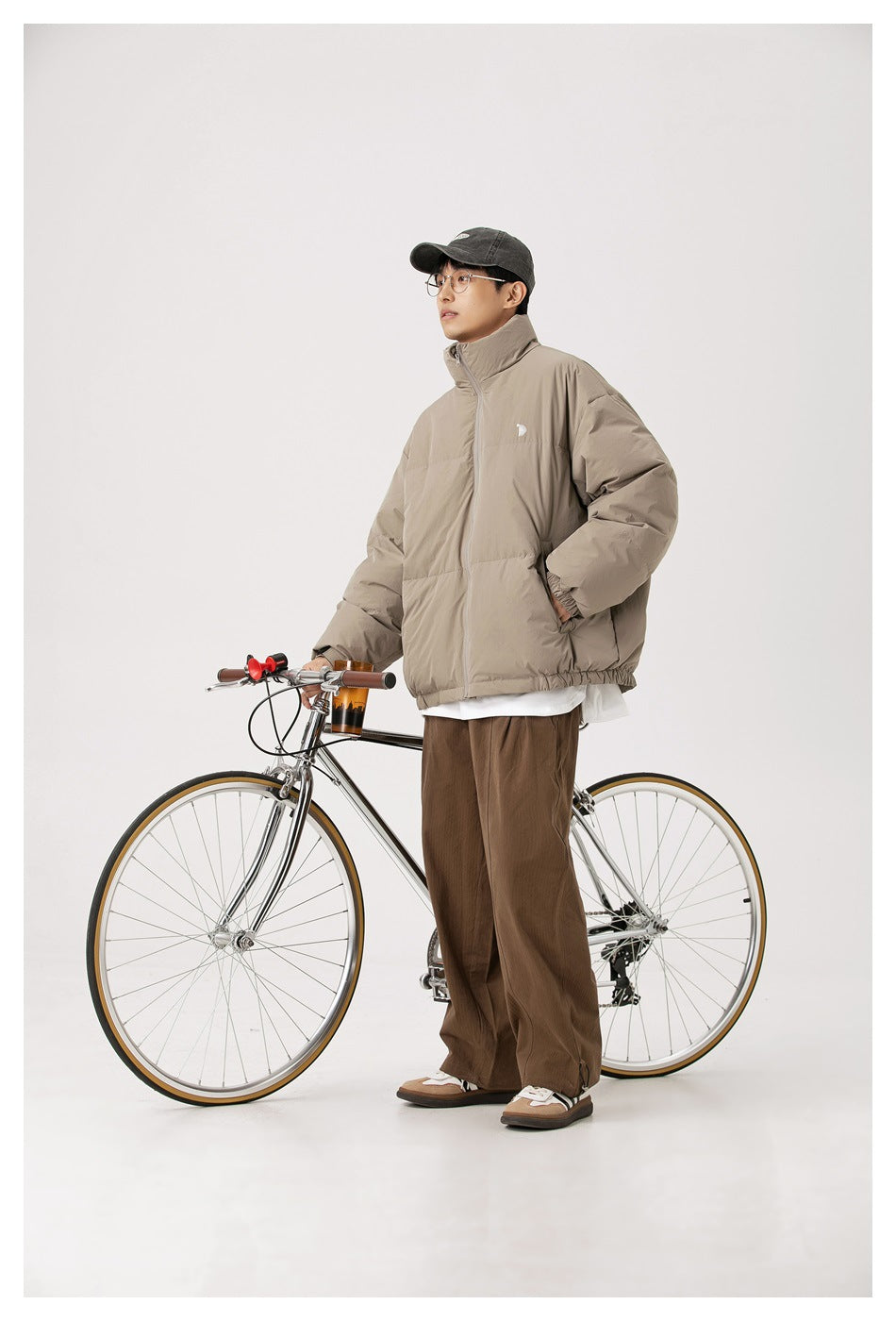 Full-length model wearing a taupe puffer jacket beside a bicycle.