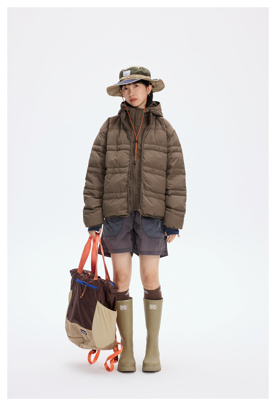 Full-length front view of a model in a brown hooded puffer jacket, gray shorts, and outdoor accessories.