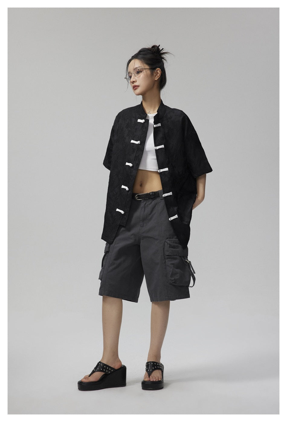 Model wearing a black short-sleeve jacket with white frog closures, styled with dark cargo shorts.