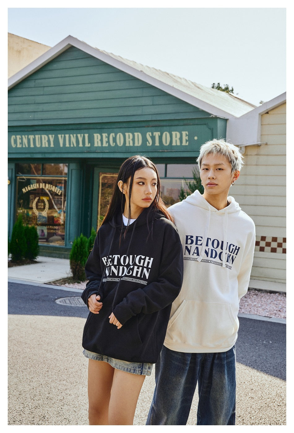 Two models wear black and white graphic pullover hoodies, shown from the front with chest lettering and relaxed fits.