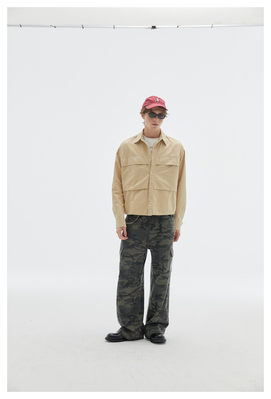 Model wearing a beige button-up overshirt with large front pockets and loose dark camouflage cargo pants.