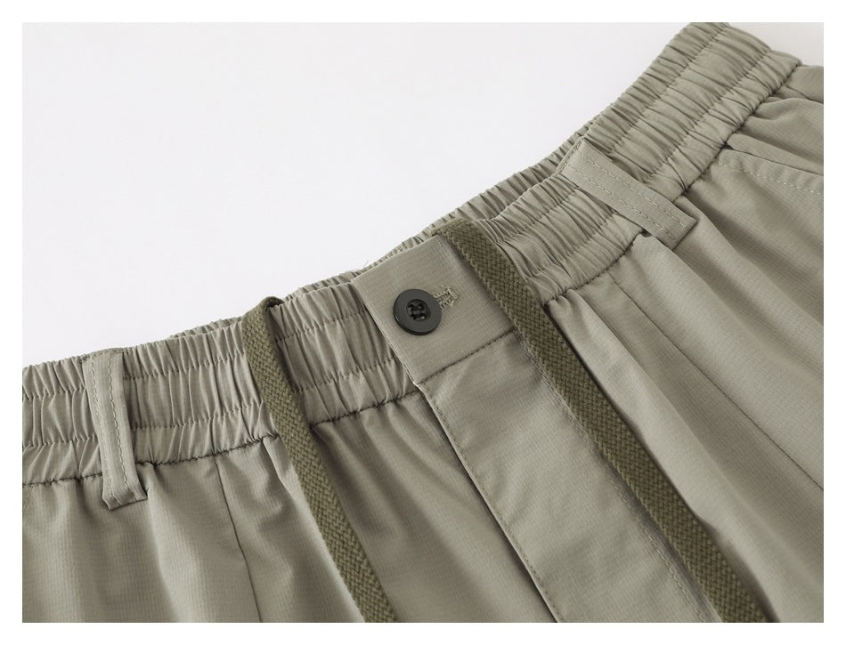Close-up of light gray-green shorts showing the gathered waistband, belt loops, button closure, and textured drawstring.