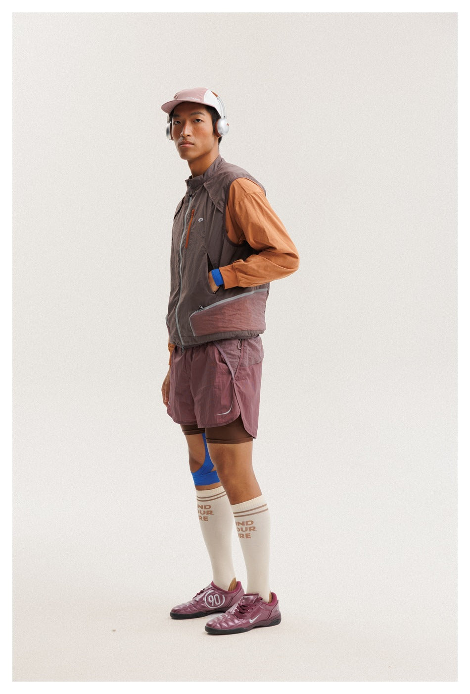 Side view of a man wearing a taupe sleeveless vest over an orange top and mauve shorts.
