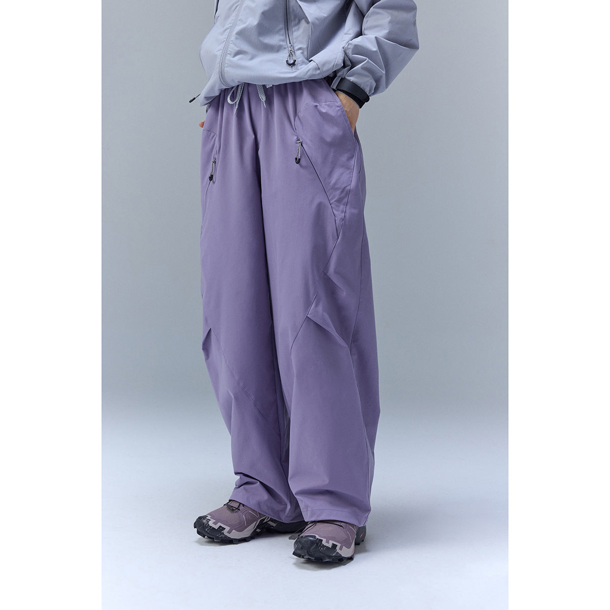 Cropped model wearing loose lavender outdoor pants with a drawstring waist and zippered pockets.