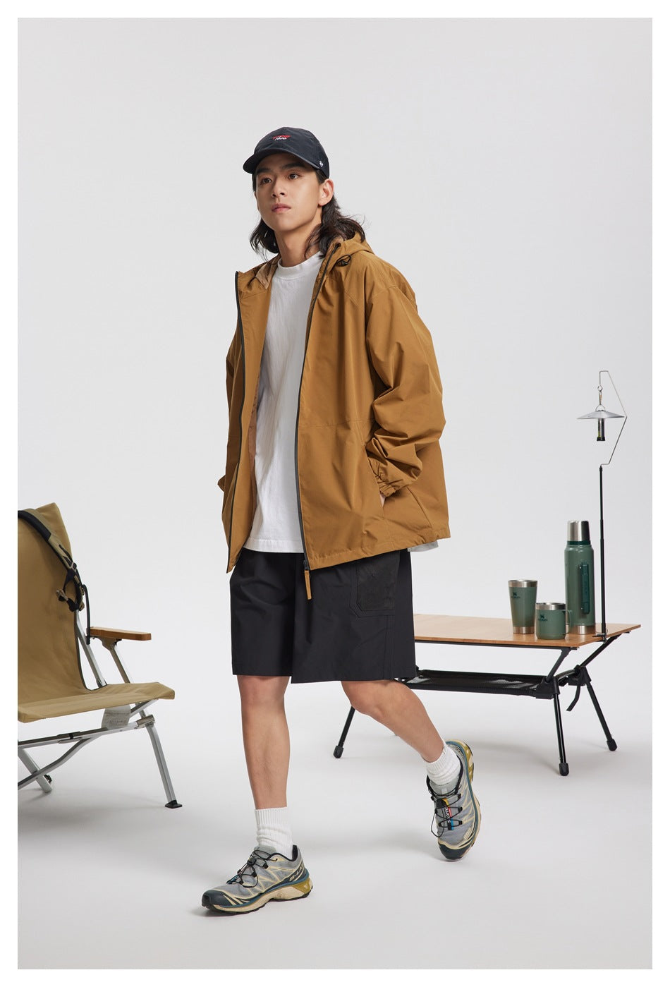 Model wearing an open mustard-brown hooded jacket over a white shirt, with black shorts.