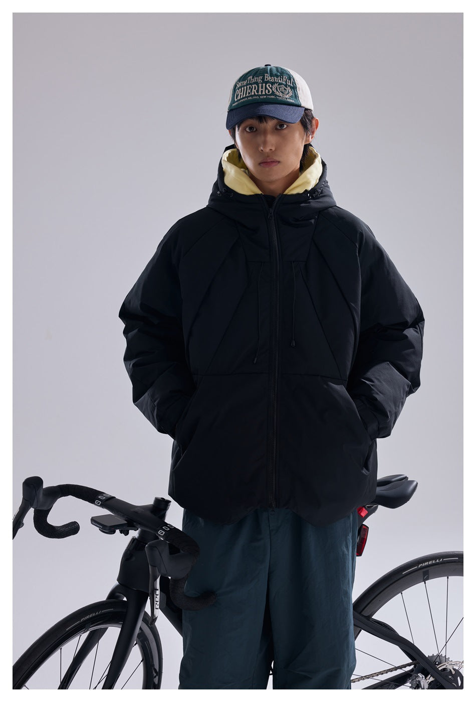 Model wearing a zipped black hooded puffer jacket over a pale yellow hoodie beside a bicycle.