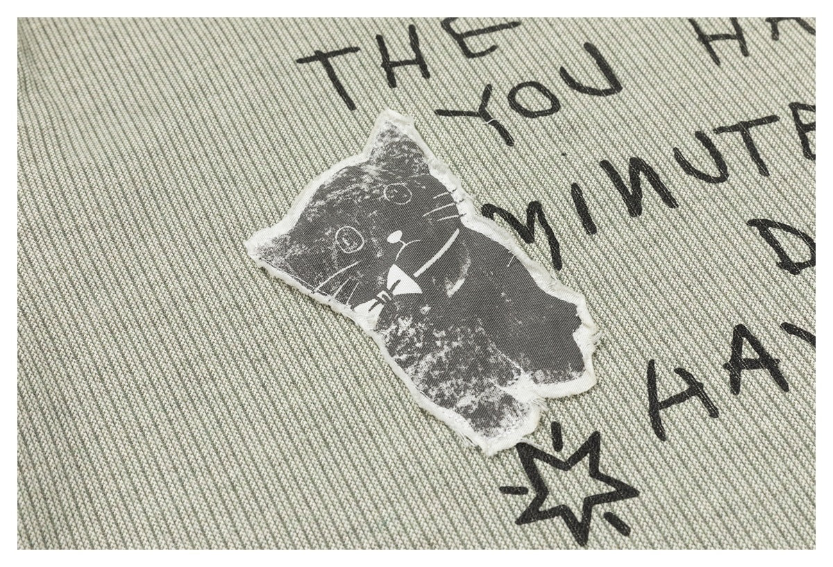 Close-up of a distressed gray cat patch and black lettering on sage-gray textured shirt fabric.