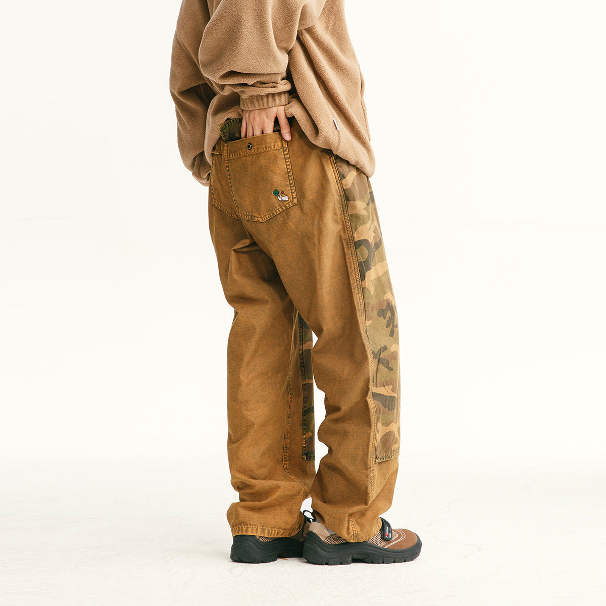 Angled rear view of a model wearing loose ochre-brown pants with camouflage side panels and back pockets.